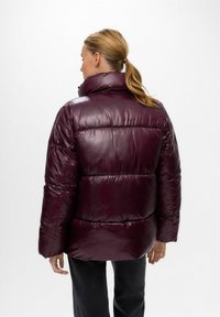 Bourgogne pufferjacka med glänsande textur, hög krage och stygnsektioner för isolering, bärs över mörka byxor.