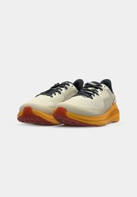 Laufschuhe mit einem hellen Cremobermaterial, schwarzen Schnürsenkeln und einer strukturierten Oberfläche. Hervorhebung einer dicken, orangen Zwischensohle und einer roten Außensohle.