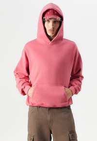 Sweat-shirt à capuche en coton rose avec une poche avant, coupe décontractée et poignets côtelés, porté sur un pantalon cargo marron.