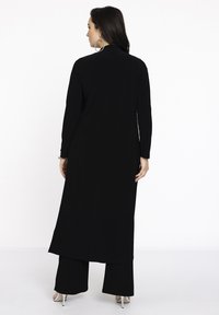 Lange schwarze Tunika mit langen Ärmeln, geradem Schnitt und Seitenschlitzen, getragen über weiten schwarzen Hosen und hohen Sandalen. Glatte Textur.