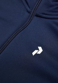 Giacca blu navy con zip e texture liscia; presenta un logo bianco sul petto e una zip frontale con un sottile tirante a contrasto.