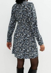 Abito nero floreale con motivi di fiori blu e bianchi, maniche lunghe, vita aderente e gonna svasata. Indossato con collant neri e stivali fino al ginocchio.