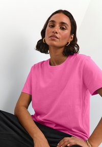 T-shirt ajusté à col rond en coton rose vif, avec des manches courtes et une texture lisse, associé à un bas sombre. Design simple.