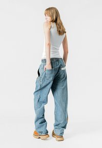 Pantalones vaqueros holgados de mezclilla azul clara con cinturilla elástica, bolsillos laterales y detalle de parche en la parte trasera. Llevados con una camiseta de tirantes blanca ajustada.