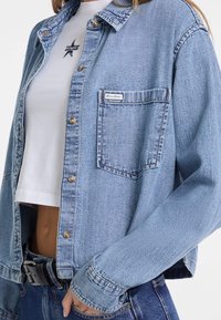 Giacca in denim azzurro chiaro con chiusura a bottone, una tasca sul petto e una vestibilità morbida. Presenta lievi segni di usura e una finitura testurizzata.