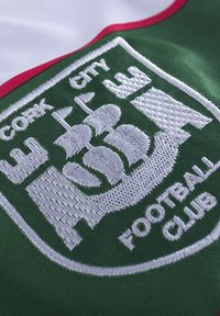 Groene stof met een wit geborduurd embleem met een schip, kasteeltorens en de tekst "Cork City Football Club" onderaan.