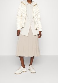 Poszewana, kremowa kurtka z kapturem, w połączeniu z jasnobeżową, plisowaną spódnicą i białymi butami sportowymi z fakturowaną podeszwą.