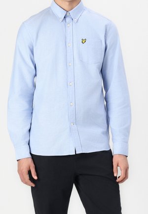 Man wearing light blue button-up shirt with a small yellow bird logo on the chest pocket and black pants, standing against white background.