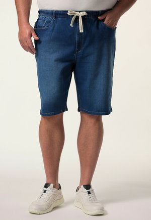 Mand iført blå denimshorts med hvid snøre og hvide sneakers, stående med hænderne i lommerne mod en ensfarvet baggrund.