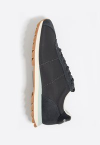 Zapatilla deportiva de tejido gris oscuro con un talón de ante texturizado, suela blanca y verde claro, y costuras decorativas a lo largo de los laterales.