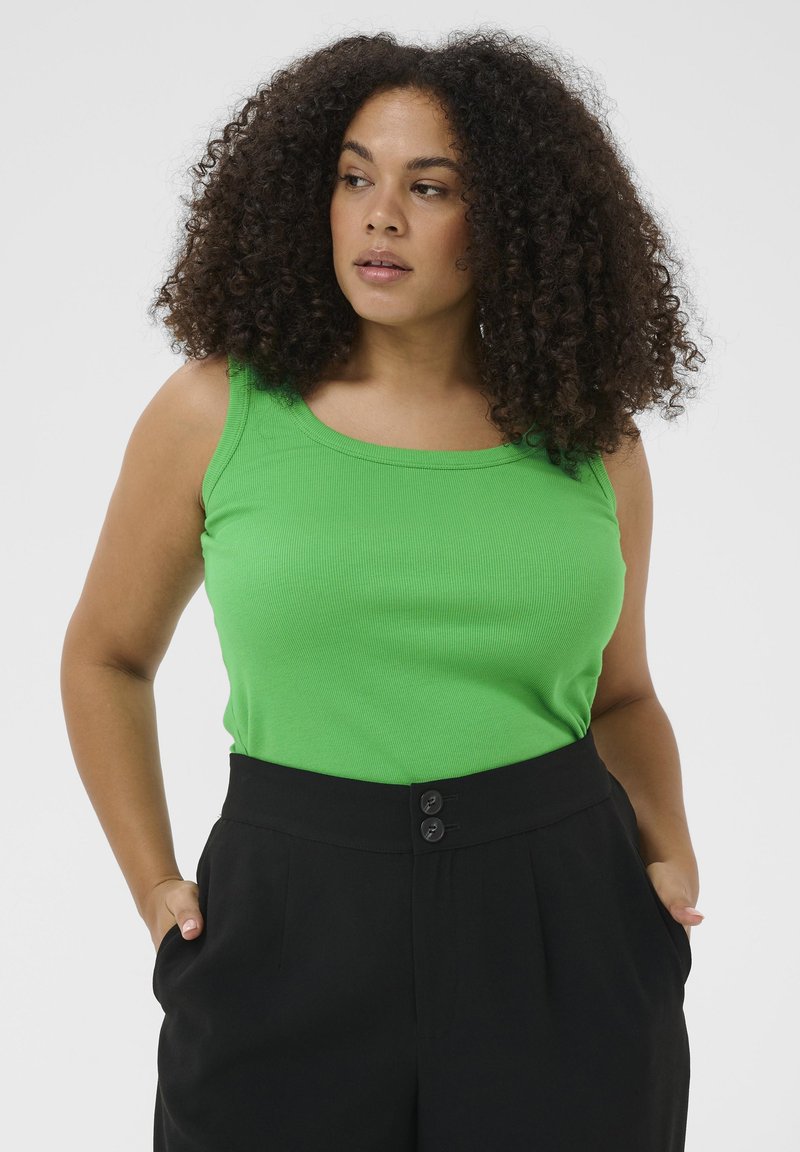 Femme aux cheveux bouclés portant un haut sans manches vert vif et un pantalon noir taille haute, les mains dans les poches, regardant sur le côté.