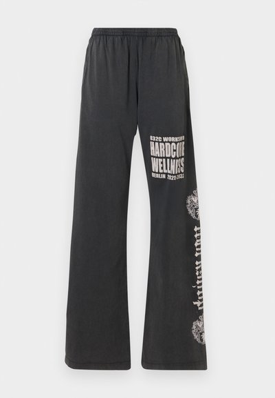 Mustad lõhevärvilised püksid elastse vöökohtaga, millel on valged graafilised printimised, mis ütlevad "032C WORKSHOP HARDCORE WELLNESS BERLIIN 2022/2023."