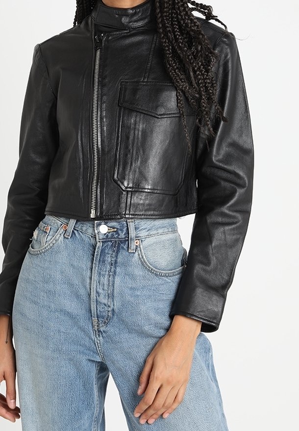 Veste cropped en cuir noir avec un col montant, fermeture éclair et une poche poitrine; portée avec un jean taille haute bleu clair.