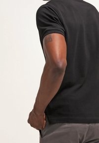 Homme portant une chemise noire à manches courtes avec un petit tatouage sur le haut du bras, debout avec la main près de sa poche.