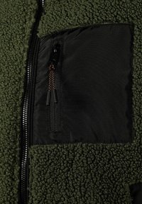 Tissu en polaire verte avec une poche zippée en nylon noire, présentant une surface texturée et un tirant de fermeture éclair orange contrastant.