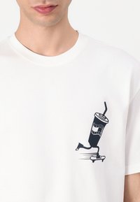 Hvid kortærmet t-shirt med et sort grafisk motiv af et drikkekrus på en skateboard, der fremhæver enkle linjer og minimalistisk design.