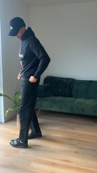 Sweatshirt preto com capuz, com um bolso frontal e cordão cinza, combinado com calças pretas e sapatos pretos, situado numa sala com iluminação suave.
