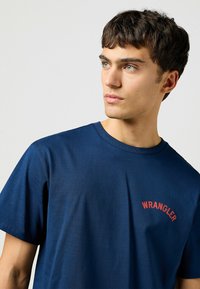 Tummansininen puuvillapaita lyhyillä hihoilla, jossa on punainen "WRANGLER"-logo rinnassa. Tasainen rakenne ja klassinen pyöreä pääntie.