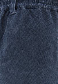 Navyblauwe corduroy stof met verticale ribbelstructuur. Beschikt over een elastische tailleband zonder extra hardware of accenten.