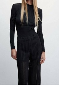 Femme portant un haut noir ajusté à manches longues avec des détails aux épaules et un pantalon noir à rayures verticales taille haute sur fond uni.