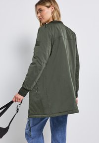 Oliivinvihreä pommijakku, jossa on ribbikujat, sivuvetoketjut ja teksturoitu kangas. Vasemmassa olkassa on logopainatus.