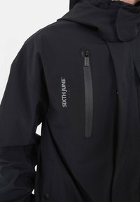 Giacca nera impermeabile con tasca frontale con zip, cappuccio regolabile e dettaglio del logo. Texture liscia con design moderno.
