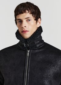 Giacca in ecopelle nera con finitura texturizzata, dotata di un colletto alto foderato con pelliccia spessa e scura e chiusura frontale con cerniera.