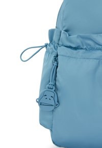 Lichtblauwe, zachte nylon tas met geplooide bovenkant, trekkoordsluiting en een textuur van een aapvormige ritsenhanger bevestigd.