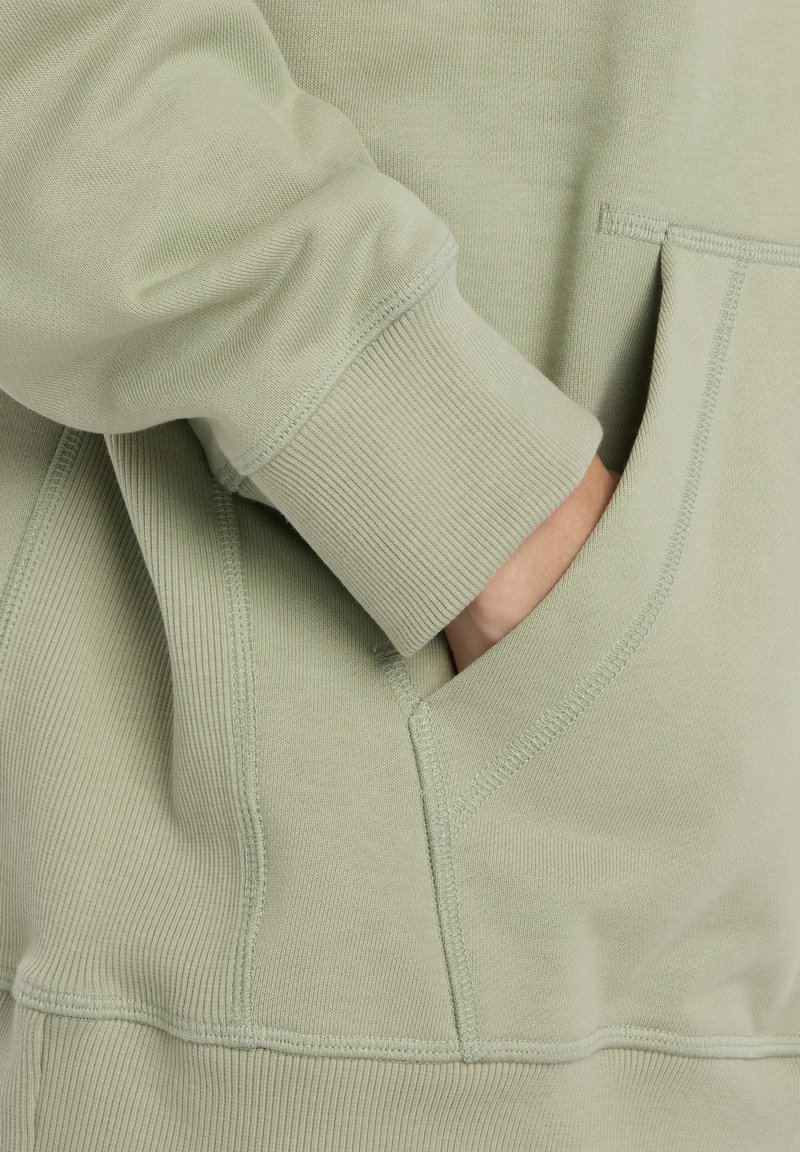 Sweat à capuche vert clair avec poignets côtelés et poche frontale, présentant des coutures plates et une texture douce. Détail sur le bras et la poche.