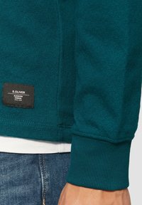 Sweatshirt teal en tissu texturé, avec un patch logo en bas et des poignets côtelés. Design classique à col rond.