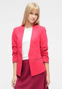 Blazer rosa acceso su misura con collo a scialle, maniche lunghe e cerniere in vita. Indossato sopra una maglietta bianca e una gonna color borgogna.