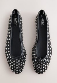 Ballerines noires avec des clous métalliques brillants couvrant la surface supérieure, dotées d'un bout rond et d'un détail de nœud noir à l'avant.