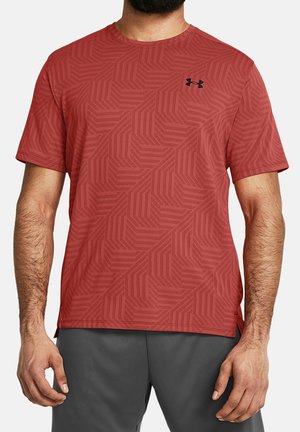 Mężczyzna w czerwonej sportowej koszulce z krótkim rękawem, ozdobionej geometrycznym wzorem i czarnym logo na piersi, w połączeniu z ciemnoszarymi spodniami.