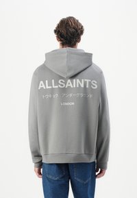 Grå hettegenser med en stor hvit "ALLSAINTS" logo på baksiden, med snorhette og ribbede mansjetter. Kombinert med blå jeans.
