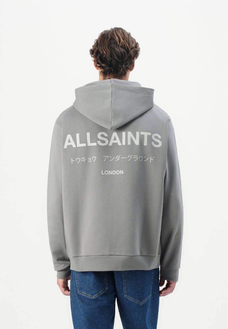 Grå hettegenser med en stor hvit "ALLSAINTS" logo på baksiden, med snorhette og ribbede mansjetter. Kombinert med blå jeans.