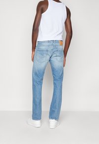 Vaqueros de mezclilla azul claro con corte recto, con un parche de cuero en la parte posterior de la cintura y bolsillos traseros clásicos, combinados con una camiseta sin mangas blanca.