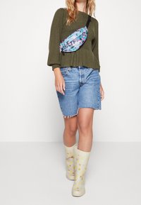 Marsupio con un motivo astratto blu e viola, maglione verde oliva testurizzato, pantaloni denim corti e stivali da pioggia a pois in un morbido giallo e beige.