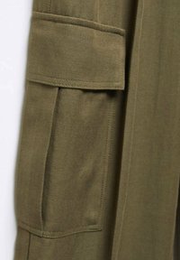 Gros plan d’un tissu vert olive avec une poche à rabat rectangulaire et des coutures visibles sur un vêtement.