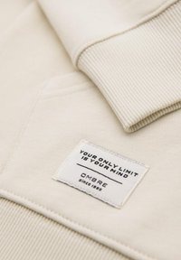 Felpa color crema con polsini a costine, dotata di un'etichetta tessuta che mostra una frase motivazionale e il nome del marchio "OMBRE, dal 1985."