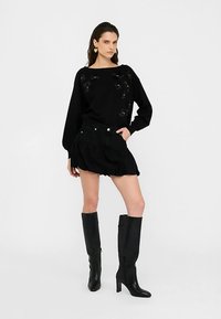 Juodas laisvo dizaino megztinis su gėlių dekoracijomis, derinamas su raukšlėtu juodu mini sijonu ir juodais batais iki kelių.