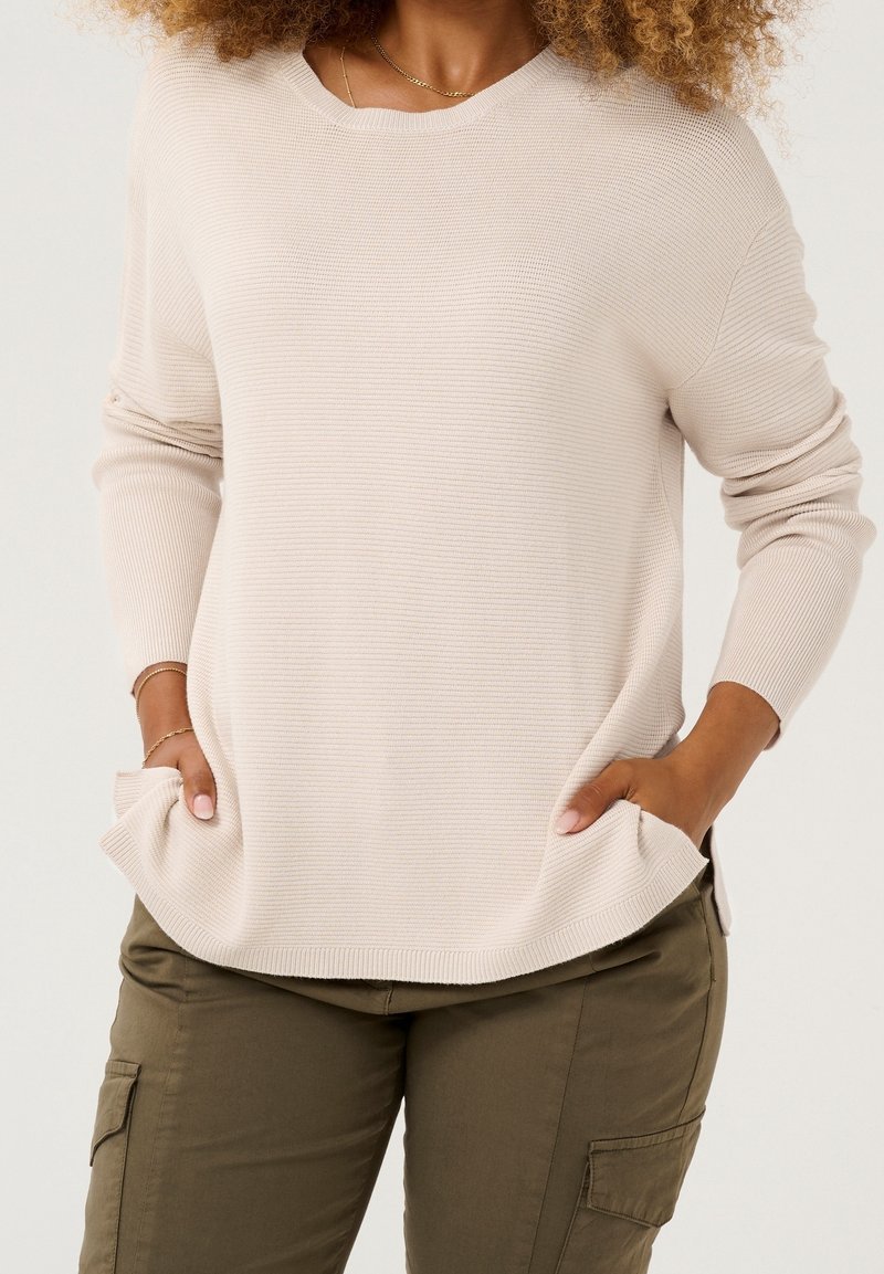 Hellbeiger gerippter Pullover mit rundem Halsausschnitt und Seitentaschen, kombiniert mit olivgrünen Cargohosen mit Seitentaschen.