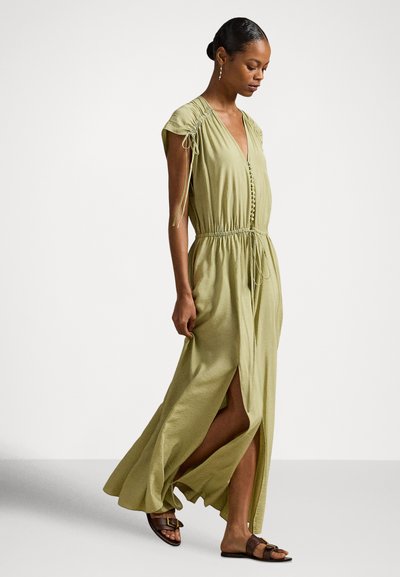 Γυναίκα που περπατά φορώντας ένα μακρύ φόρεμα σε ανοιχτό πράσινο χρώμα με μανίκια καπέλο, κουμπιά μπροστά, πλαϊνή σχισμή και καφέ ίσια σανδάλια με αγκράφες.