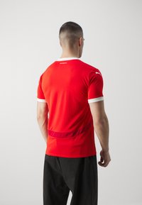 Crvena sportska majica s bijelim ovratnikom i akcentima na rukavima. Prilagodljiv dizajn izrađen od mekog materijala. Ima suptilnu zakrivljenu teksturu na leđima.