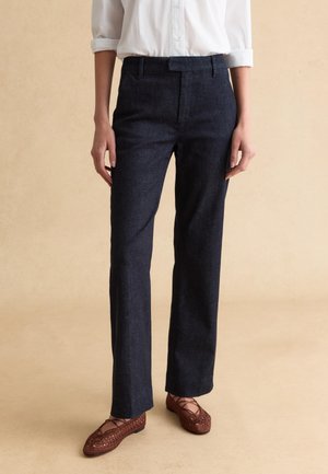 Persona con pantalones oscuros de pierna recta, camisa blanca con mangas remangadas y zapatos planos marrones tejidas, sobre un fondo beige.