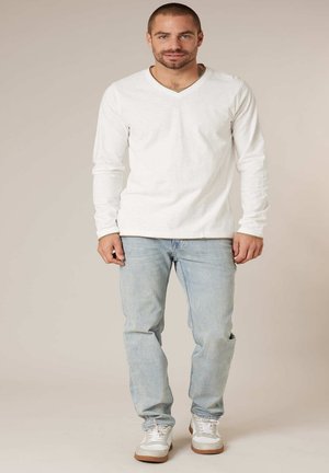 Homme debout face à l'avant, portant une chemise blanche à manches longues avec col en V, un jean bleu clair et des baskets blanches sur un fond beige.