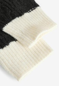 Muffole in lana lavorate a maglia con una parte superiore nera e un polsino a coste color crema. Texture morbida con fibre visibili e dettagli di cucitura a contrasto.