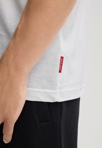 Biała bawełniana koszulka z widoczną czerwoną metką "JOOP! JEANS" po boku, z krótkimi rękawami i luźnym krojem. Czarne spodnie częściowo widoczne.