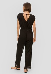 Zwarte jumpsuit met korte mouwen, V-rug en een cinched taille. Gemaakt van lichte stof, met een zijslit en strikdetail. Te zien zijn slippers.