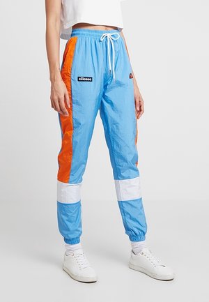 Osoba nosząca niebieskie spodnie dresowe z pomarańczowymi paskami po bokach, białymi opaskami przy łydkach, białym sznurkiem i białymi trampkami.