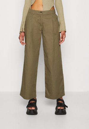 Persoon draagt een olijfgroene broek met brede pijpen en zwarte sandalen met open tenen, staand tegen een effen witte achtergrond.